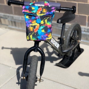 May include: Black balance bike with a colorful splatter print handlebar bag. The bag has a blue button closure.