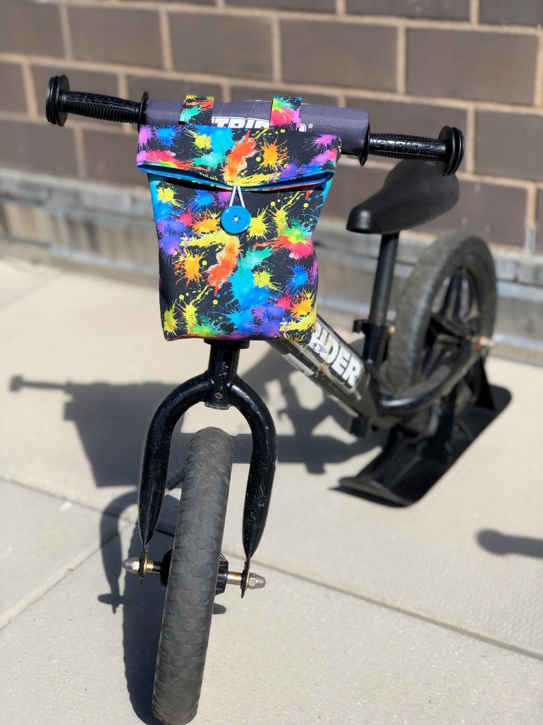 May include: Black balance bike with a colorful splatter print handlebar bag. The bag has a blue button closure.