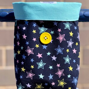 K&ouml;nnte beinhalten: Eine schwarze Stofftasche mit t&uuml;rkisfarbenem Oberteil und einem gelben Knopf. Die Tasche hat ein Sternenmuster in verschiedenen Farben, darunter rosa, blau, gelb und gr&uuml;n.