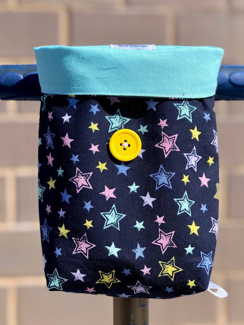 K&ouml;nnte beinhalten: Eine schwarze Stofftasche mit t&uuml;rkisfarbenem Oberteil und einem gelben Knopf. Die Tasche hat ein Sternenmuster in verschiedenen Farben, darunter rosa, blau, gelb und gr&uuml;n.
