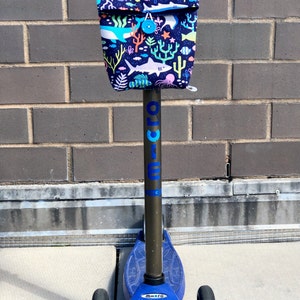 Peut inclure: Une trottinette bleue avec un guidon noir et un sac en tissu attach&eacute; au guidon. Le sac a un imprim&eacute; color&eacute; de cr&eacute;atures marines, y compris des requins, des poissons et des coraux. La trottinette a le mot "micro" imprim&eacute; sur le c&ocirc;t&eacute;.