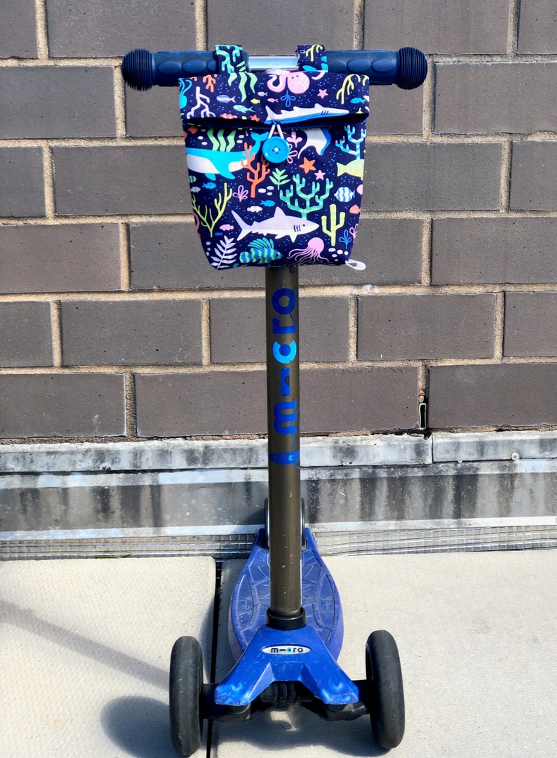 Peut inclure: Une trottinette bleue avec un guidon noir et un sac en tissu attach&eacute; au guidon. Le sac a un imprim&eacute; color&eacute; de cr&eacute;atures marines, y compris des requins, des poissons et des coraux. La trottinette a le mot "micro" imprim&eacute; sur le c&ocirc;t&eacute;.