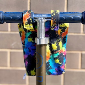 May include: A black fabric bag with colorful paint splatter design attached to a scooter handlebar. The bag has two straps that attach to the handlebar.