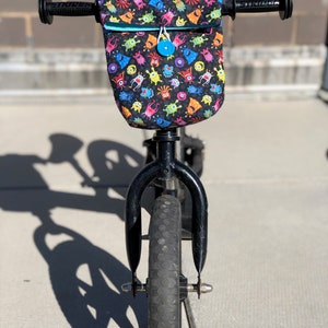 May include: A black balance bike with a front handlebar bag featuring a colorful monster print. The bag has a blue zipper and a blue button.