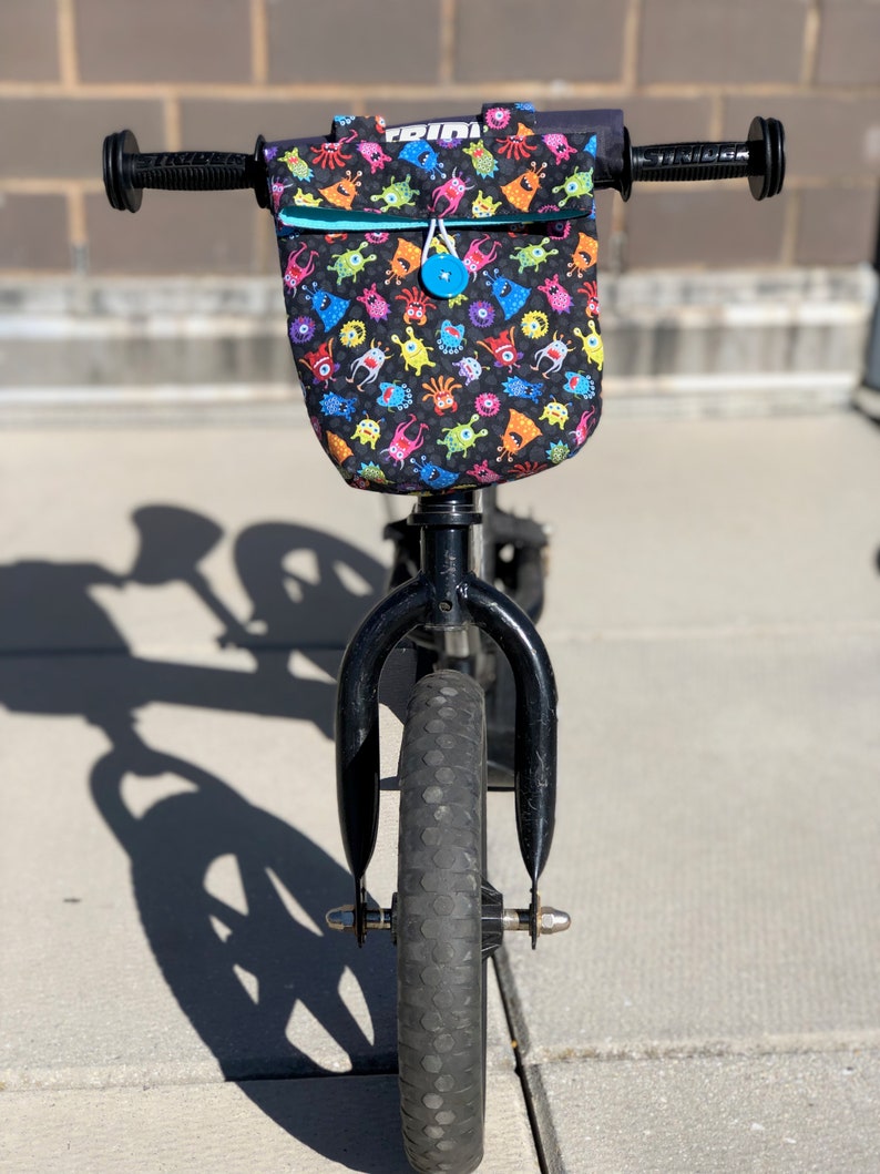 May include: A black balance bike with a front handlebar bag featuring a colorful monster print. The bag has a blue zipper and a blue button.