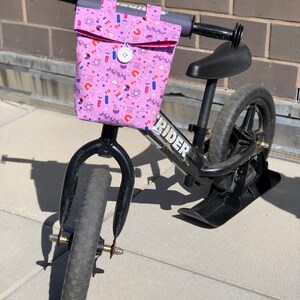 May include: A black balance bike with a pink handlebar bag featuring a science-themed print. The bag has a flap with a button closure. The bike has a black seat and black wheels.