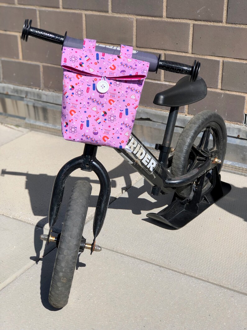 May include: A black balance bike with a pink handlebar bag featuring a science-themed print. The bag has a flap with a button closure. The bike has a black seat and black wheels.