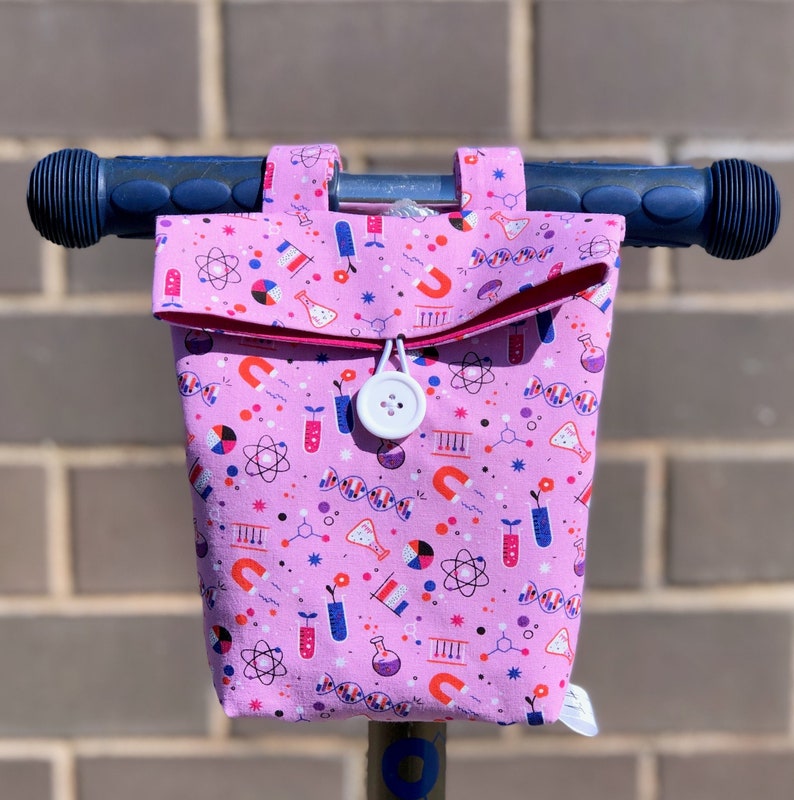 May include: Pink fabric handlebar bag with a science theme print. The bag has a white button closure and a pink zipper. The print features atoms, test tubes, DNA strands, and magnets.