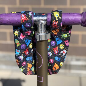 May include: Purple scooter handlebars with black fabric handlebar covers featuring a colorful monster print.
