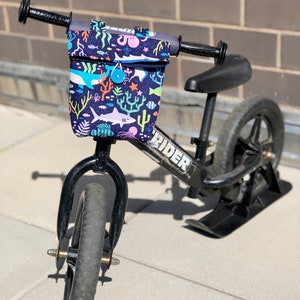 Peut inclure: Un v&eacute;lo d'&eacute;quilibre noir avec un sac de guidon imprim&eacute; de motifs marins color&eacute;s. Le sac a un fond bleu avec des poissons color&eacute;s, des coraux et une pieuvre. Le v&eacute;lo a une selle noire et des roues noires. Le v&eacute;lo porte l'inscription "RIDER" sur le cadre.