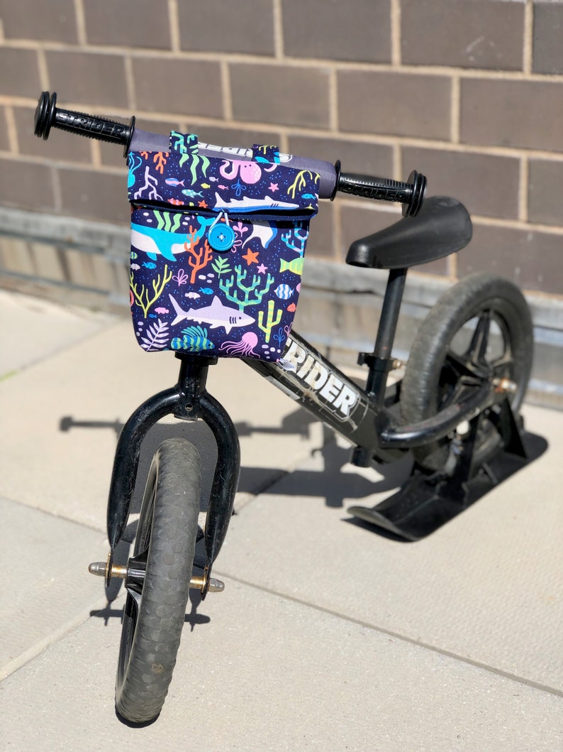 Peut inclure: Un v&eacute;lo d'&eacute;quilibre noir avec un sac de guidon imprim&eacute; de motifs marins color&eacute;s. Le sac a un fond bleu avec des poissons color&eacute;s, des coraux et une pieuvre. Le v&eacute;lo a une selle noire et des roues noires. Le v&eacute;lo porte l'inscription "RIDER" sur le cadre.