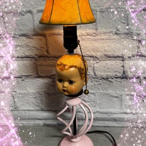 May include: A table lamp featuring a doll head base with painted facial features and light brown hair. The lamp has a pink spiral metal stand and a circular base. An orange fabric lampshade sits atop the head, controlled by a gold pull chain.