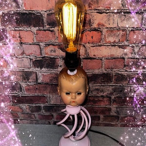 May include: A table lamp featuring a vintage doll head base with a light pink spiral stand and a round base. An Edison-style light bulb with a visible glowing filament is mounted directly on top of the doll head, emitting a warm yellow light.