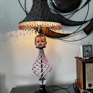 Może przedstawiać: Lampa stołowa z czarnym abażurem wykończonym frędzlami z bursztynowych koralików. Podstawa składa się z głowy lalki umieszczonej nad różową, spiralną konstrukcją, przezroczystej szklanej kuli oraz czarnej, okrągłej podstawy.