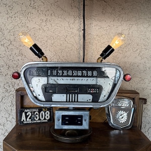 May include: A unique lamp crafted from vintage car parts. The lamp features a speedometer, two light bulbs, and a license plate that reads "ARIZONA A2-308 GRAND CANYON STATE". The lamp is a creative blend of industrial and retro design.