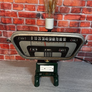 May include: A table lamp constructed from a vintage automotive dashboard gauge cluster featuring a speedometer, temperature, fuel, oil, and battery indicators. The lamp includes a clear Edison-style light bulb and a dark green base with a white electrical outlet.