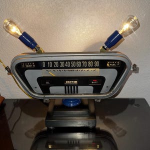 May include: A table lamp constructed from a vintage automotive dashboard gauge cluster. The unit features a speedometer marked 0 to 90, oil and battery indicators, and two blue-based light bulbs extending from the top. It sits on a black base.