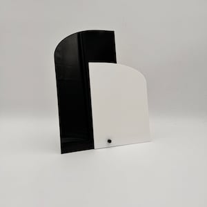 Może przedstawiać: Abstrakcyjny dekor ścienny w kolorze czarnym i białym wykonany z dwóch akrylowych elementów. Większy element jest czarny i ma kształt łuku. Mniejszy element jest biały i ma kształt półkola. Oba elementy są połączone ze sobą u góry.