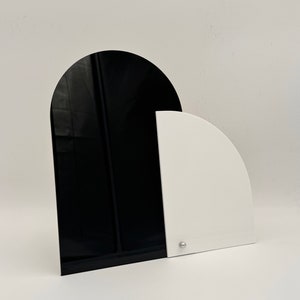 May include: Two black and white arch-shaped pieces of acrylic stand on a white surface. The larger piece is black and the smaller piece is white. The pieces are joined together at the top.