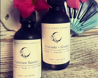 May include: Two black glass bottles with white labels. The bottles are labeled "Tahitian Sunset" and "Avocado + Ginger". Both bottles are room and car sprays. The bottles are sitting on a wooden surface with a pink flower.