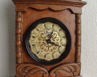 May include: A wooden clock with a carved frame and a black face with Roman numerals. The clock face has a gold background with a decorative design and the word "Miera" in the center.