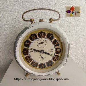 May include: White alarm clock with a gold face and black Roman numeral markings. The clock has a black hand and a small, round, black alarm clock face with a white hand. The clock face says "Tornado K Loud Alarm" and "Jerge".