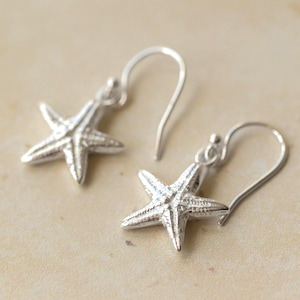 May include: A pair of silver starfish earrings. Each earring has a detailed starfish charm hanging from a curved ear wire. The earrings are shown against a neutral background, emphasising their metallic shine.