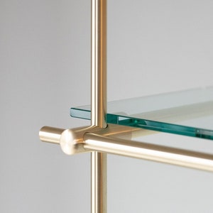 May include: Close-up of a brass shelf unit with a glass shelf. The shelf is supported by a vertical brass rod and a horizontal brass rod. The glass shelf is clear and has a slight green tint.
