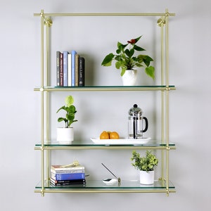 May include: A gold metal shelf unit with three glass shelves. The shelves are decorated with books, plants, and a French press coffee maker.