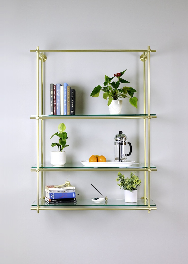 May include: A gold metal shelf unit with three glass shelves. The shelves are decorated with books, plants, and a French press coffee maker.