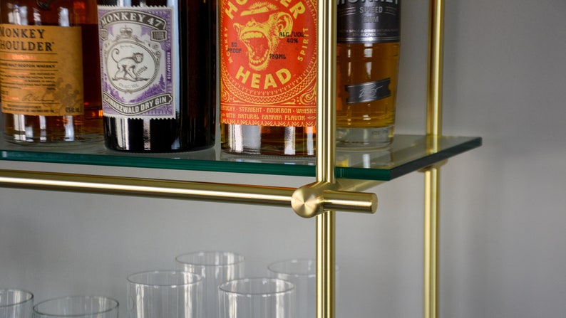 May include: A gold metal bar cart with two glass shelves. The top shelf has four bottles of liquor, including Monkey Shoulder, Monkey 47, and a bottle with a red label that says "Hot Head". The bottom shelf has eight empty glasses.