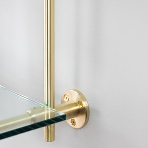 May include: A gold metal shelf bracket with a round base and three screws. The bracket is attached to a glass shelf.
