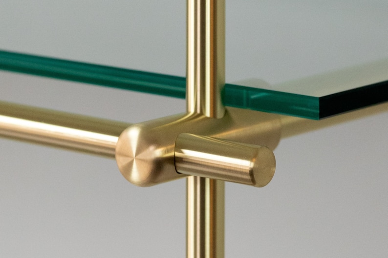 May include: Close-up of a brass shelf bracket with a glass shelf. The bracket has a round, polished finish and is attached to a vertical brass rod.