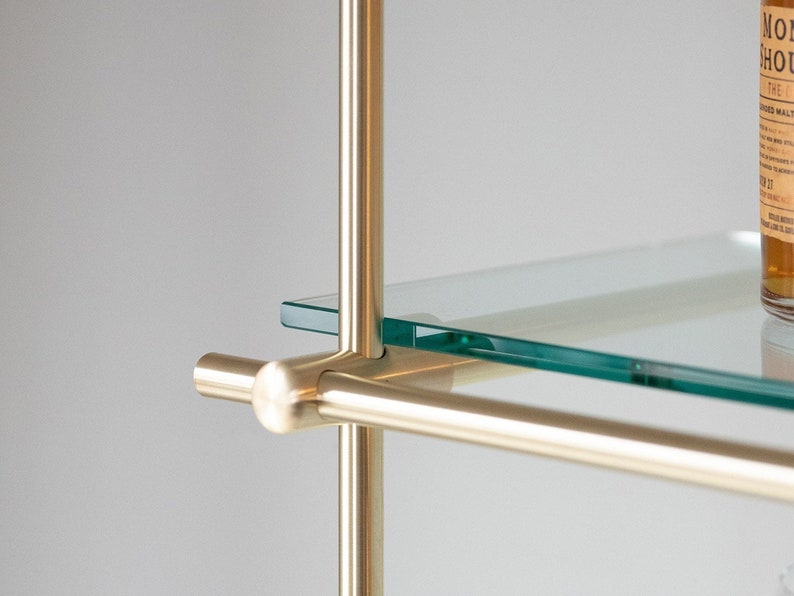 May include: Close-up of a brass shelf unit with a glass shelf. The shelf is supported by a vertical brass rod and a horizontal brass rod. The glass shelf is clear and has a slight green tint.