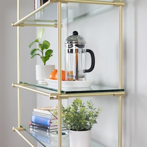 May include: A gold metal shelf unit with four glass shelves. The shelves are filled with various items, including plants, books, and a French press coffee maker.