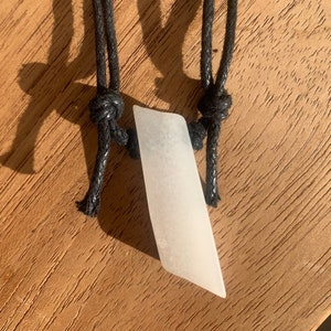 May include: A white, opaque, smooth, and slightly curved pendant hangs from a black braided cord. The pendant is shaped like a long, thin, pointed leaf.
