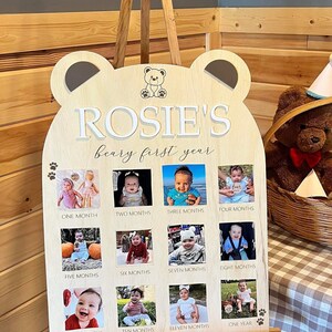 Może przedstawiać: Drewniany znak w kształcie niedźwiedzia z tekstem "ROSIE'S beary first year" i 12 miejscami na zdjęcia na pierwszy rok dziecka. Znak stoi na sztaludze i ma brązowo-biały kratkowany obrus z tyłu.