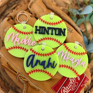 May include: A collection of softball-themed keychains. Each keychain is a neon green circle with red stitching, resembling a softball. Names are displayed in various colors: pink, blue, and black. The keychains rest on a brown leather softball glove.