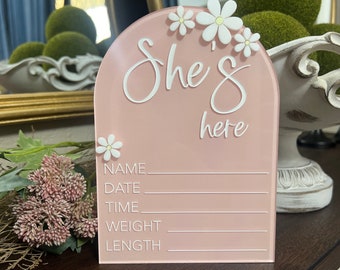 May include: Pink acrylic sign with a white arch and the text "She's here" in white script. The sign has white daisy flowers and blank lines for name, date, time, weight, and length.