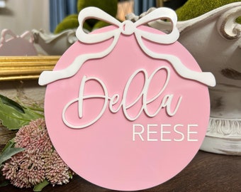 May include: Pink round ornament with a white bow and the name "Della Reese" written in white cursive lettering.