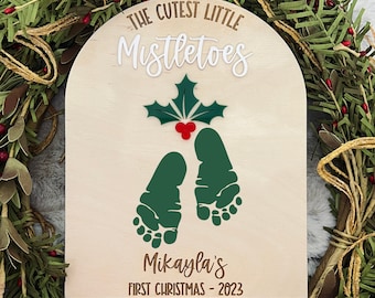 Puede incluir: Un letrero de madera con el texto "The Cutest Little Mistletoes" y una hoja de acebo verde con bayas rojas. Debajo del texto hay dos huellas verdes y el texto "Mikayla's First Christmas - 2023".