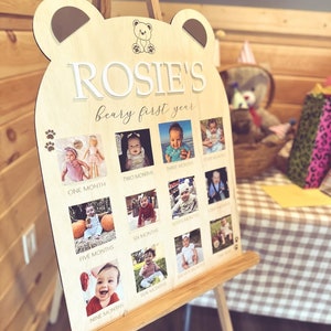 Puede incluir: Un letrero de madera con forma de oso con el texto "ROSIE'S beary first year" y fotos de una ni&ntilde;a a diferentes edades, de un mes a once meses. El letrero est&aacute; en un caballete.