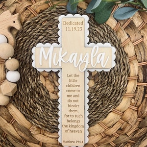 May include: A wooden cross with a white scalloped border. The cross is decorated with the name "Mikayla" and the date "11.19.23". The inscription "Dedicated" is above the name. The cross also features a bible verse: "Let the little children come to me and do not hinder them, for to such belongs the kingdom of heaven. Matthew 19:14."