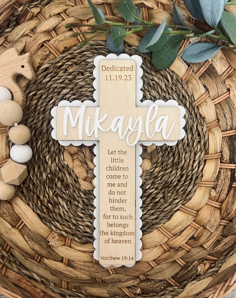 May include: A wooden cross with a white scalloped border. The cross is decorated with the name "Mikayla" and the date "11.19.23". The inscription "Dedicated" is above the name. The cross also features a bible verse: "Let the little children come to me and do not hinder them, for to such belongs the kingdom of heaven. Matthew 19:14."