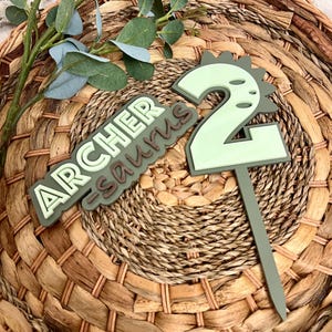Puede incluir: Topper de pastel de dinosaurio verde y marr&oacute;n con el texto "Archer -saurus" y el n&uacute;mero "2" en &eacute;l.