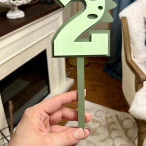 Puede incluir: Topper de pastel verde n&uacute;mero dos con un dise&ntilde;o de dinosaurio. El n&uacute;mero est&aacute; hecho de cartulina gruesa y tiene un palo verde unido a la parte posterior.