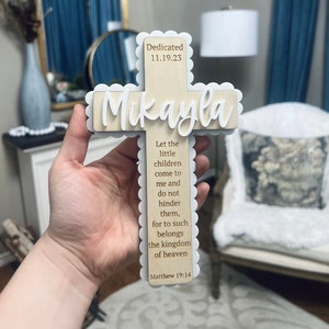 May include: A wooden cross with a white scalloped border. The cross is decorated with the name "Mikayla" and the date "11.19.23". The cross also features a bible verse from Matthew 19:14, "Let the little children come to me and do not hinder them, for to such belongs the kingdom of heaven."