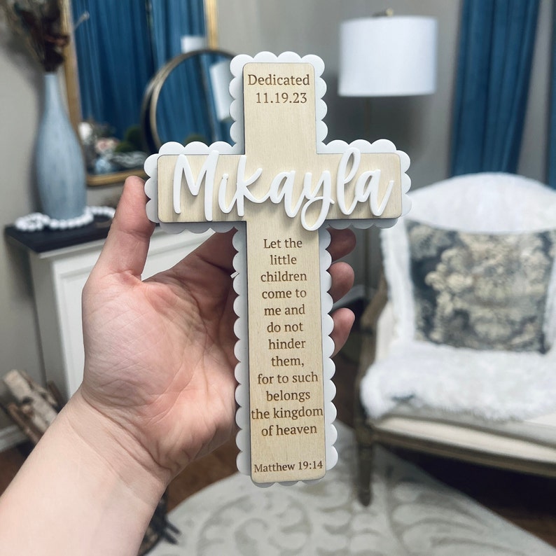 May include: A wooden cross with a white scalloped border. The cross is decorated with the name "Mikayla" and the date "11.19.23". The cross also features a bible verse from Matthew 19:14, "Let the little children come to me and do not hinder them, for to such belongs the kingdom of heaven."