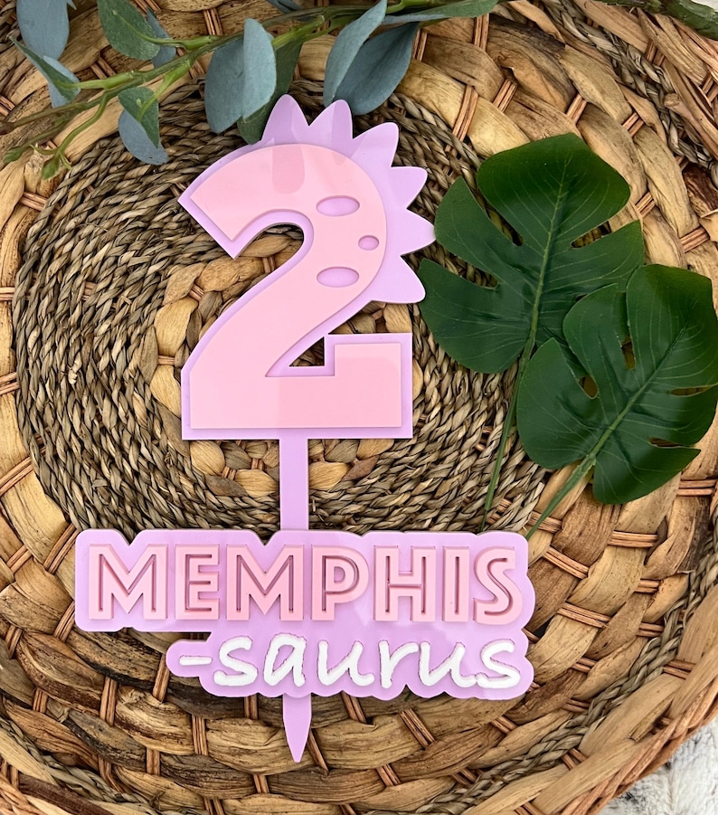 Puede incluir: Topper de pastel de dinosaurio rosa con el n&uacute;mero 2 y el nombre "Memphis-saurus" en letras blancas.