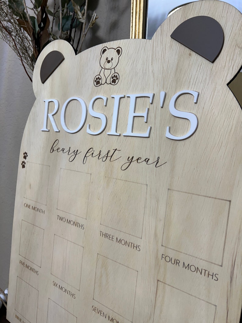 Puede incluir: Un gr&aacute;fico de crecimiento de madera en forma de oso con el nombre "ROSIE'S" y el texto "beary first year". El gr&aacute;fico tiene espacios para fotos o pegatinas para marcar cada mes del primer a&ntilde;o.